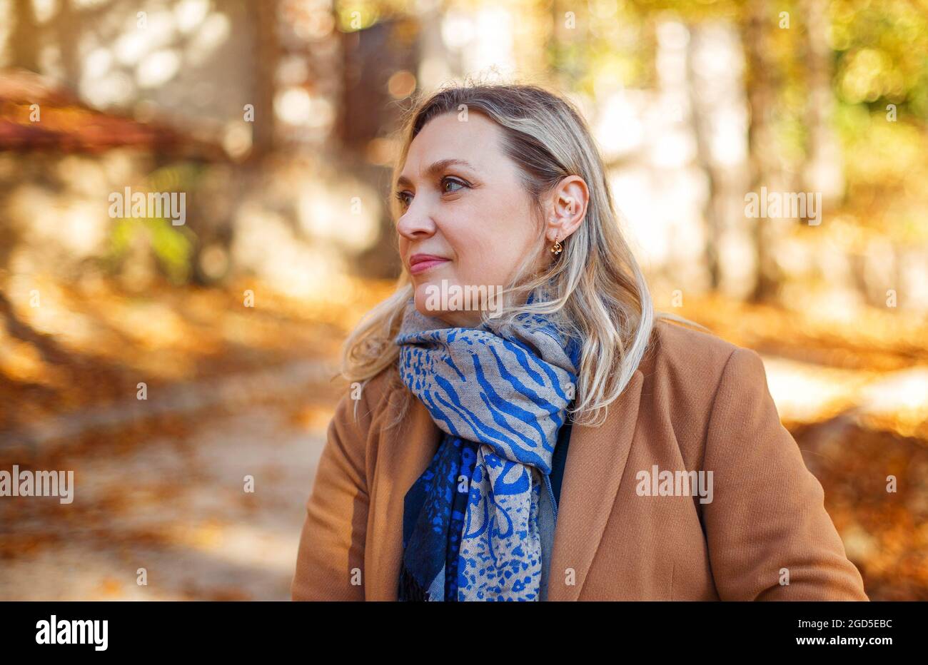 Felice donna di mezza età godendo l'autunno fuori, vestito con abiti casual e cappotto beige in piedi nel parco giallo, guardando da parte e sorridente allegro Foto Stock