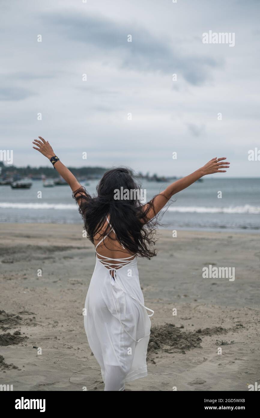 Bella donna in abito bianco a piedi sulla spiaggia. Mani laterali. Vista posteriore. Sfondo del mare. Capelli neri lunghi ricci. Vento nei capelli. Foto Stock