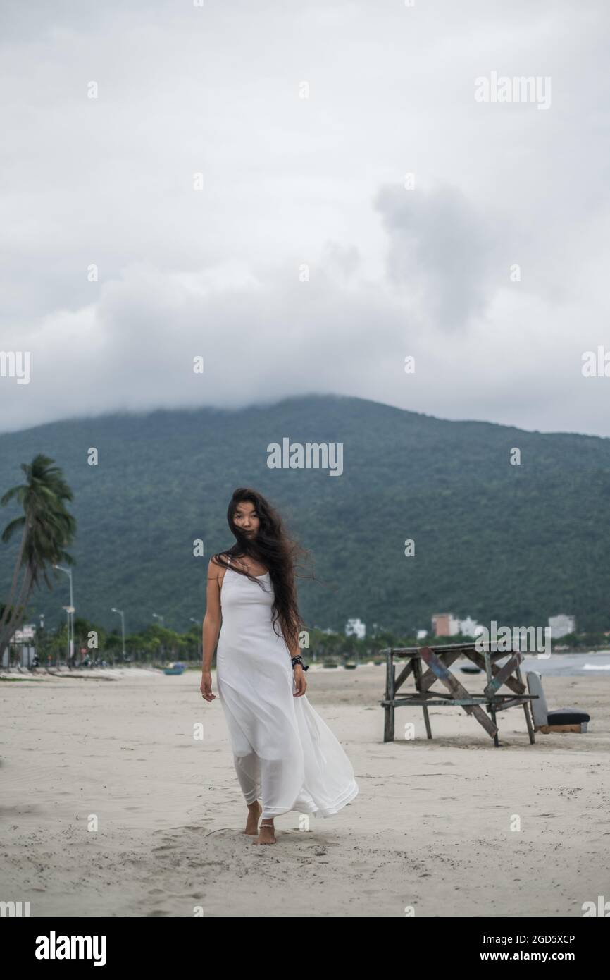 Affascinante giovane donna mongolo in abito bianco camminando sulla spiaggia. Sfondo della montagna. Capelli neri lunghi ricci. Foto di alta qualità Foto Stock