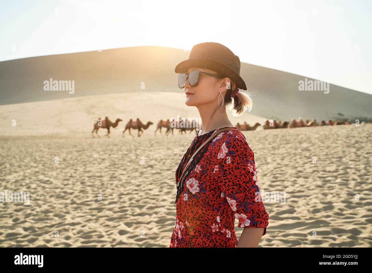 donna asiatica in abito rosso che guarda la vista nel deserto con caravan di cammelli e enormi dune di sabbia sullo sfondo Foto Stock