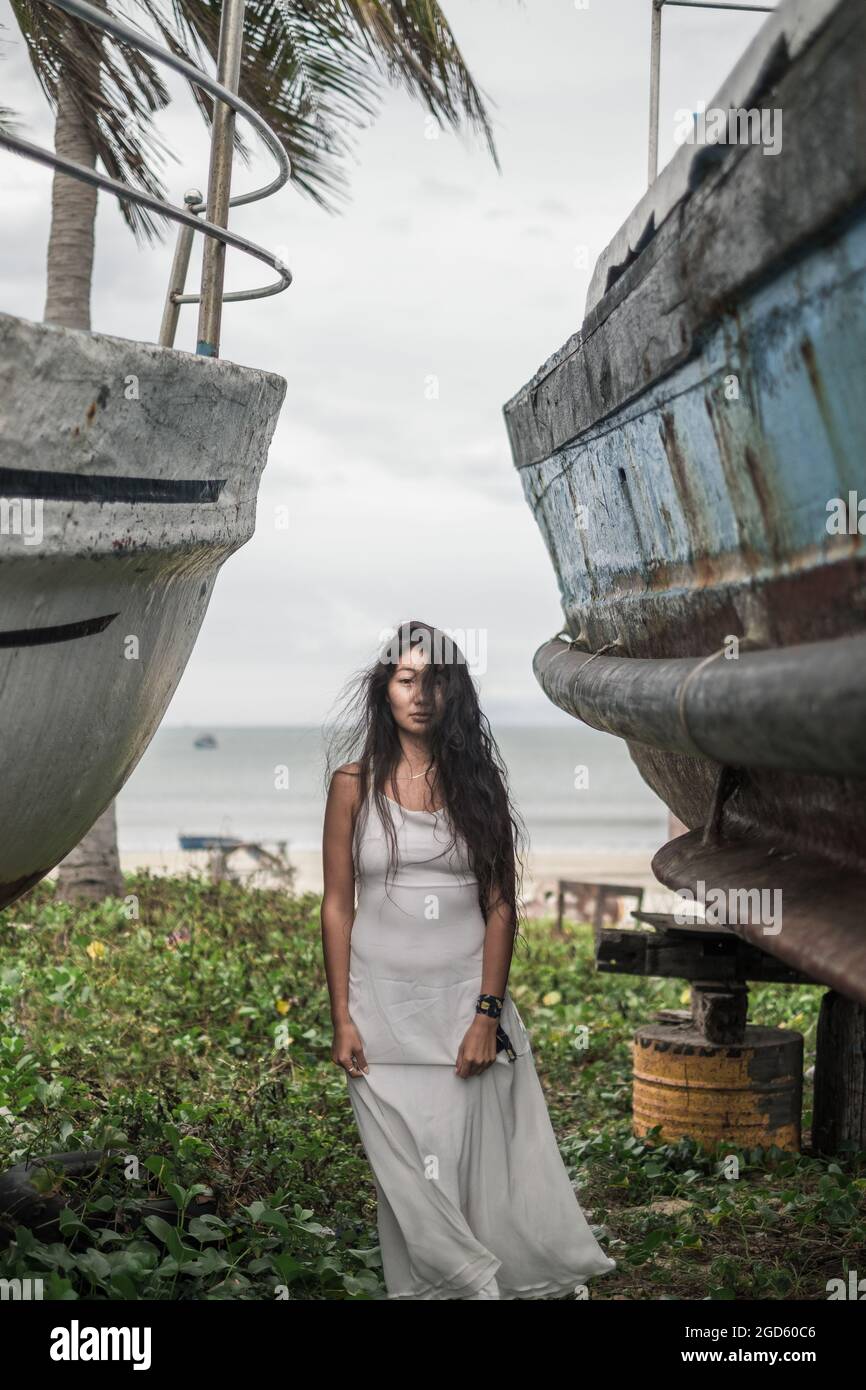 Affascinante giovane donna asiatica in grigio brillante vestito in piedi tra due vecchie navi di legno. Capelli neri lunghi ricci. Foto romantica. Giornata ventosa sulla spiaggia. Foto Stock