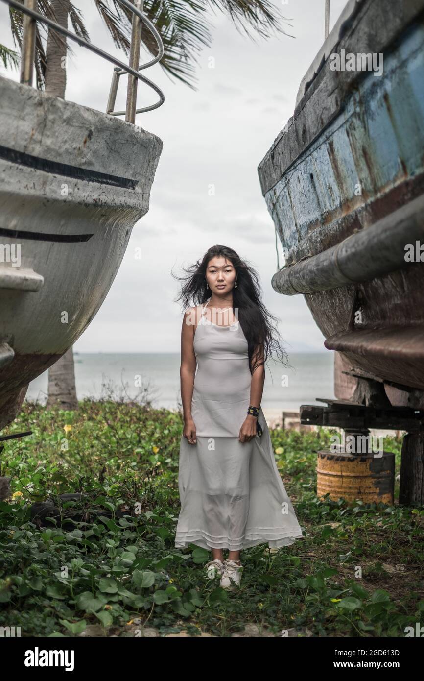 Affascinante giovane donna mongolo in abito grigio brillante in piedi tra due vecchie navi di legno. Capelli ricci. Foto romantica. Giornata ventosa. Foto di alta qualità Foto Stock