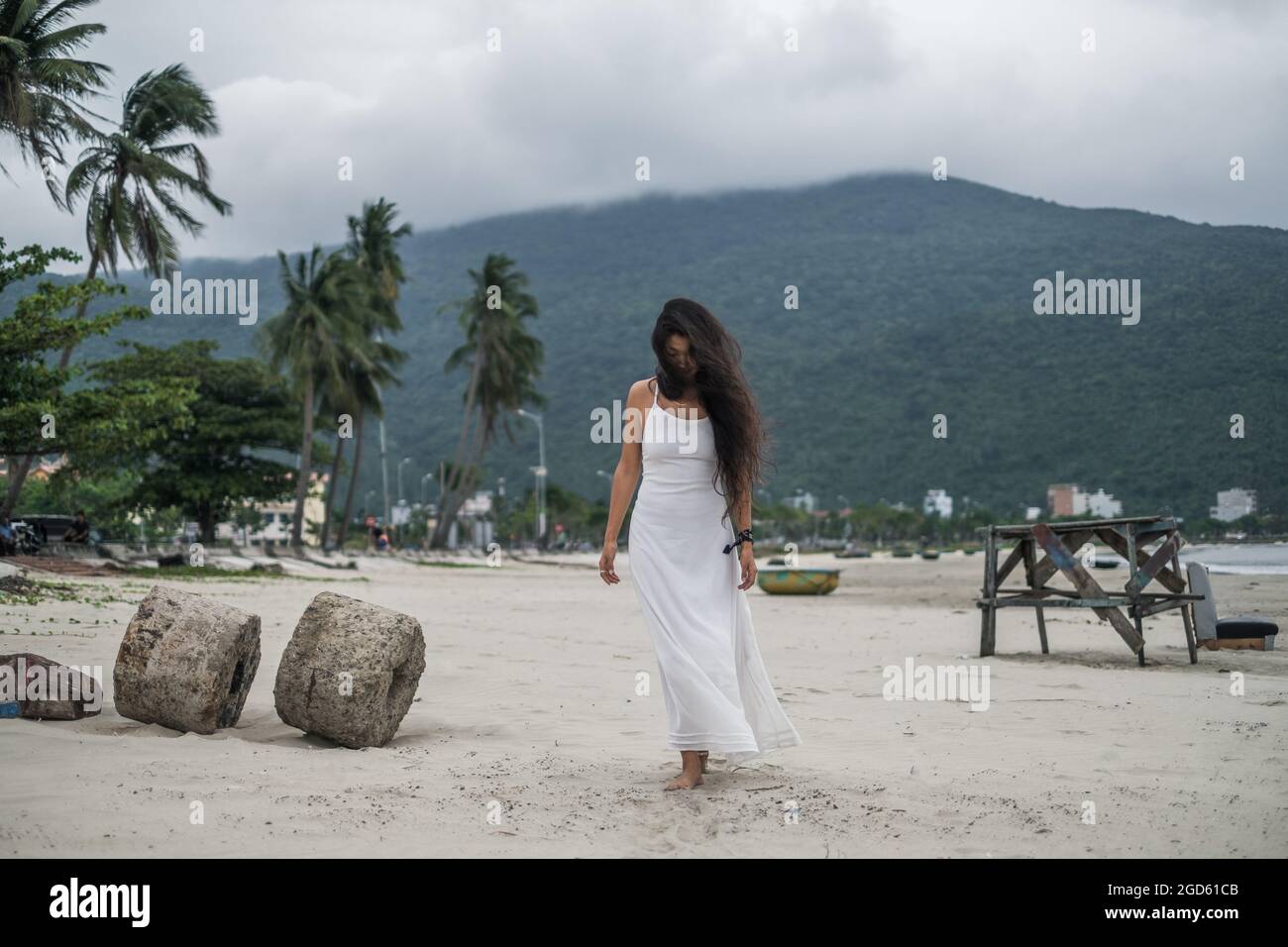 Affascinante giovane donna in abito bianco camminando sulla spiaggia e guardando verso il basso. Sfondo della montagna. Capelli neri lunghi ricci. Vento nei capelli. Foto Stock