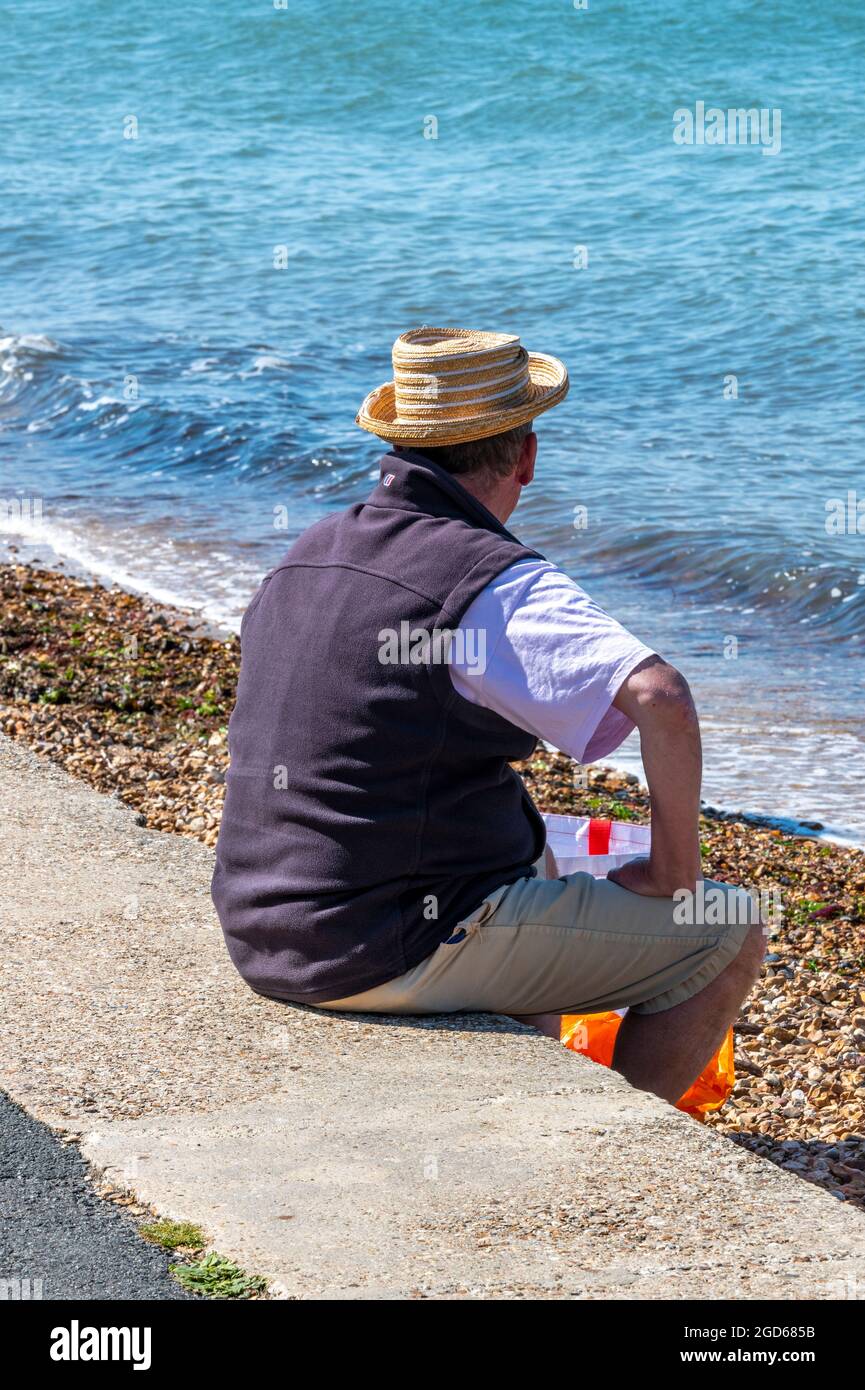 uomo seduto su una parete di mare che guarda verso il mare, uomo che  indossa cappello seduto sulla spiaggia, uomo che indossa cappello estivo,  uomo seduto sulla parete di fronte al mare