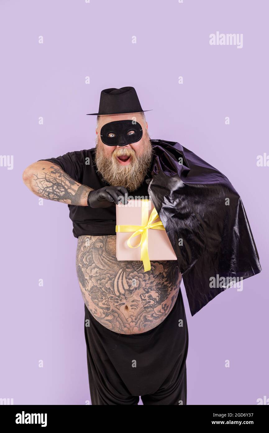 L'uomo eccitato con il peso in costume di Zorro nasconde la scatola regalo  dietro il capo su sfondo viola Foto stock - Alamy
