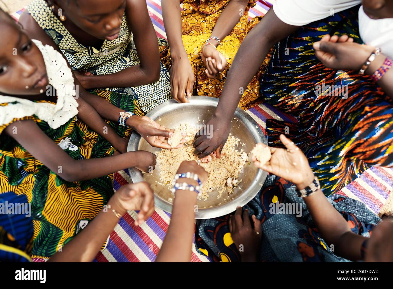 Gruppo di ragazze africane nere che si siedono su un tappetino, dividendo il loro pasto frugale, mangiando con le mani da un grande piatto di metallo Foto Stock