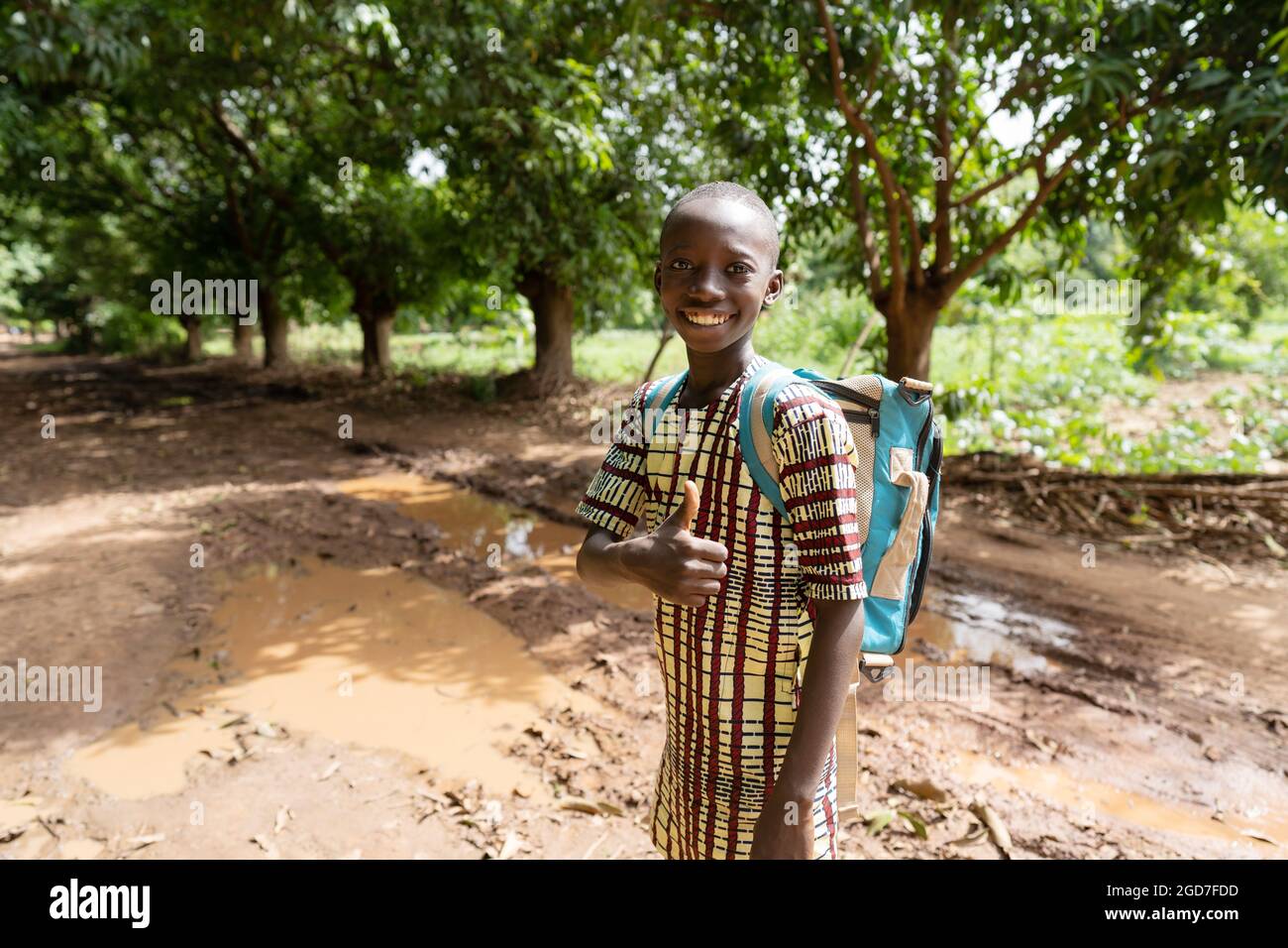 Sorridente studente africano nero sulla sua strada di casa con un sorriso felice sul viso e pollice in piedi nel fango dopo una tempesta stagionale Foto Stock