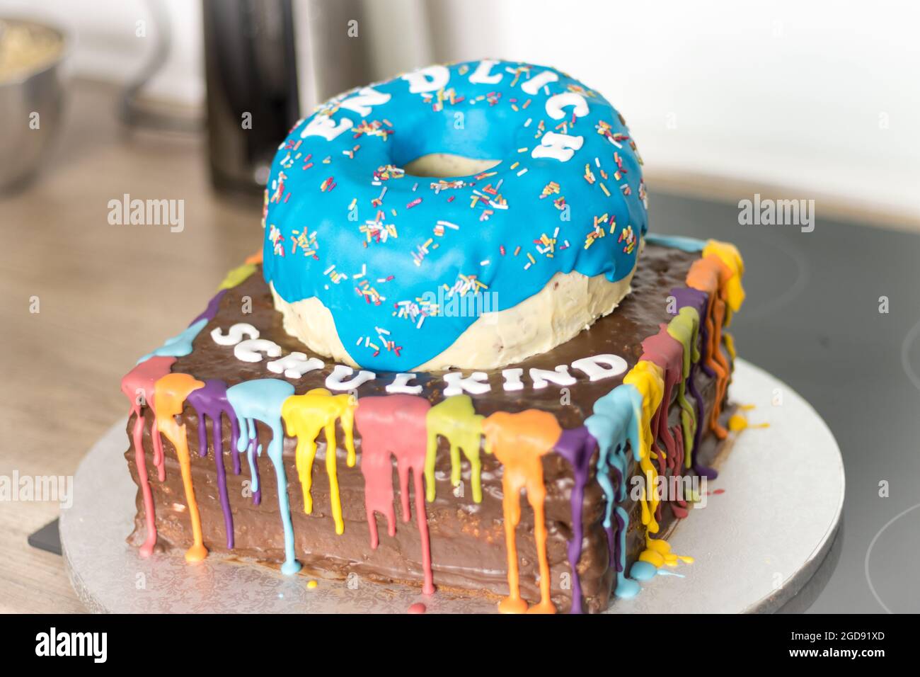 Una torta creativa con una ciambella blu sopra una colorata base quadrata ricoperta di gocciolatoio, che presenta un design vivace e divertente per un'occasione speciale. Foto Stock