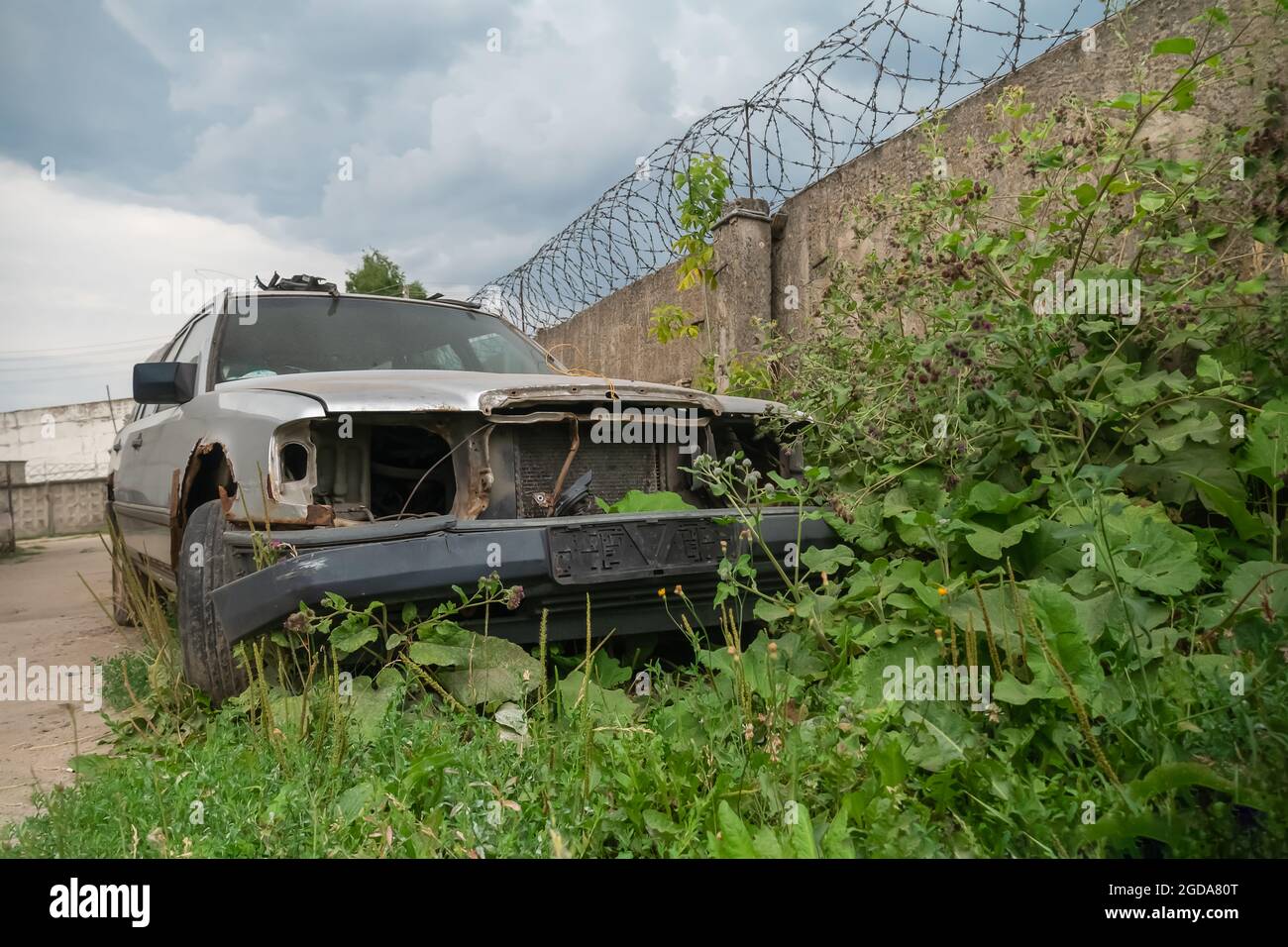 Vecchia auto grigia arrugginita senza fari vicino a una recinzione in cemento con filo spinato Foto Stock