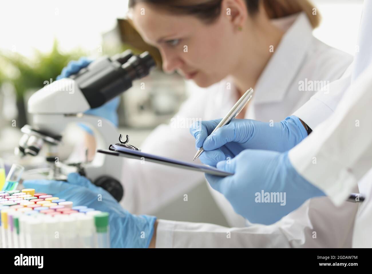 Donna farmacia che guarda attraverso il microscopio in laboratorio. Assistente che scrive i dati su carta Foto Stock