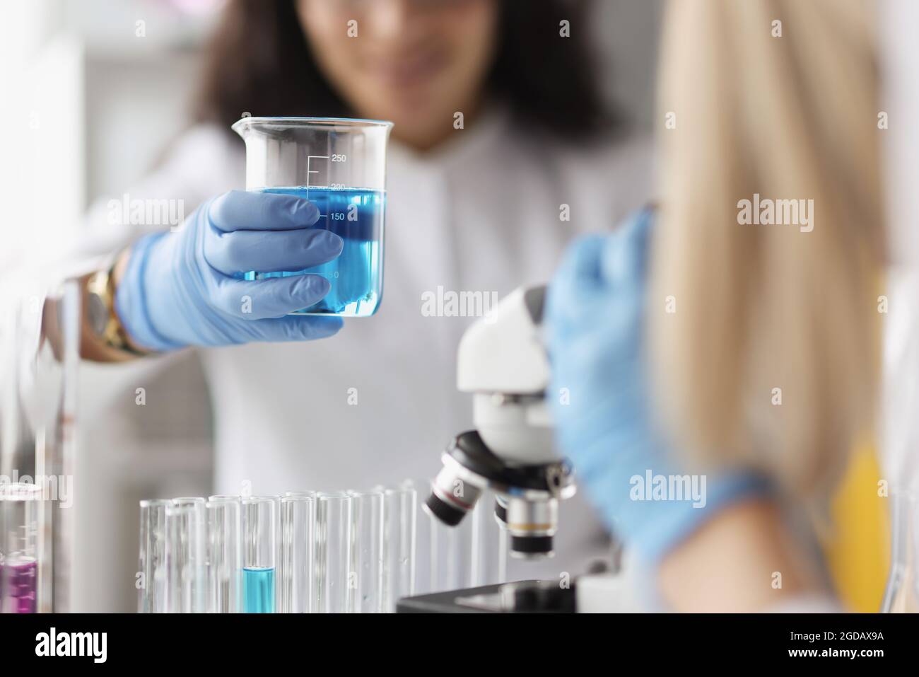 Donna chimico che tiene becher di vetro con liquido blu davanti al microscopio in laboratorio Foto Stock