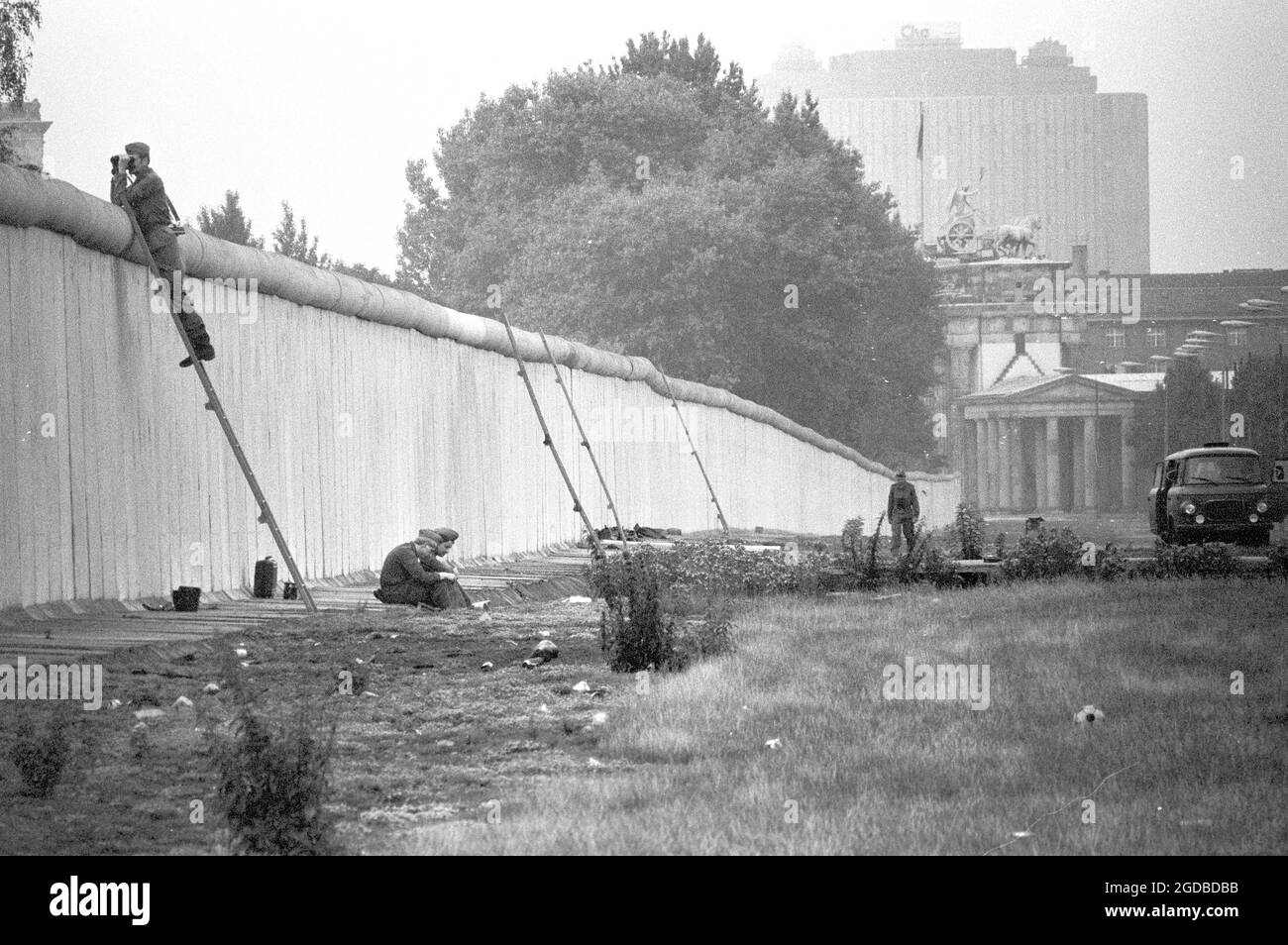 28 maggio 1988, Berlino: Le guardie di frontiera della RDT si siedono dietro il muro di Berlino, mentre un soldato su una scala osserva la situazione nel Triangolo di Lenne sul lato ovest di Berlino. Il Triangolo di Lenne era occupato all'epoca in protesta contro la costruzione della superstrada voluta qui dal Senato. Quando l'area fu bonta il 1° luglio 1988, più di 180 dei quadranti 'scapparono' sul Muro a Berlino Est, che lasciarono ancora un po' più tardi attraverso i soliti valichi di frontiera. La foto è stata scattata attraverso una piccola fessura nel Muro all'altezza dell'edificio Martin Gropius. La porta di Brandeburgo An Foto Stock