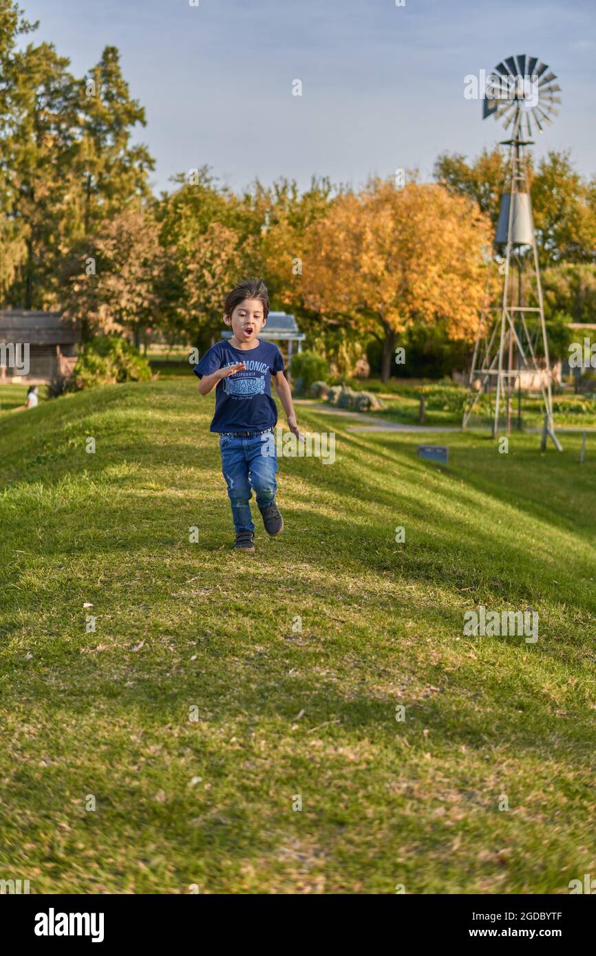 ragazzino bruna che corre su una collina e dietro di lui è un mulino a vento in autunno nella foresta. verticale Foto Stock