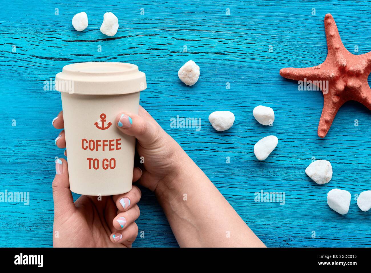 Il testo del giorno del caffè sulla tazza di caffè di bambù per andare nelle mani della donna. Testo bianco su sfondo di legno turchese con pietre bianche e stelle marine. Disposizione piatta, parte superiore Foto Stock