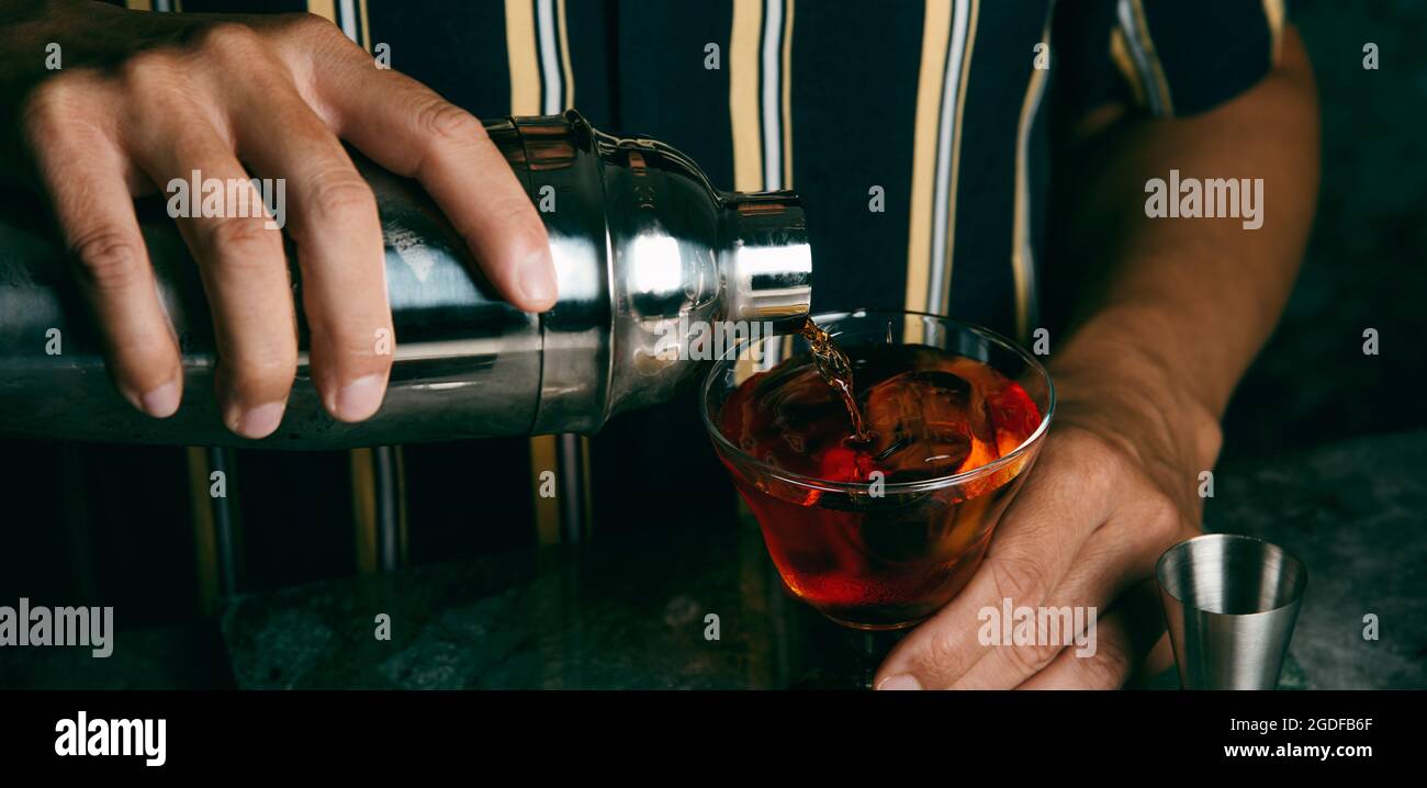 un giovane caucasico, indossando una camicia a righe, versa un cocktail, da uno shaker metallico, in un bicchiere, in un formato panoramico da utilizzare come banner web o heade Foto Stock