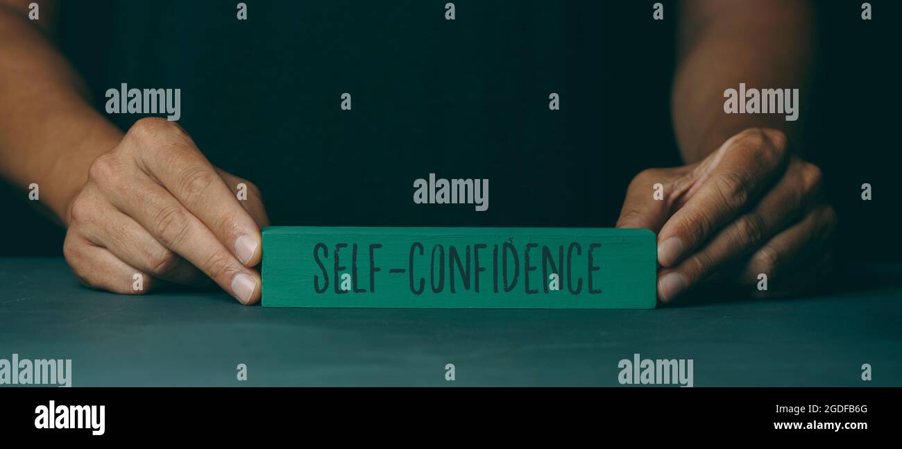 un uomo caucasico mostra un blocco di costruzione verde con il testo auto-confidenza scritto in esso, su una superficie di grigio scuro, in un formato panoramico da utilizzare come web Foto Stock