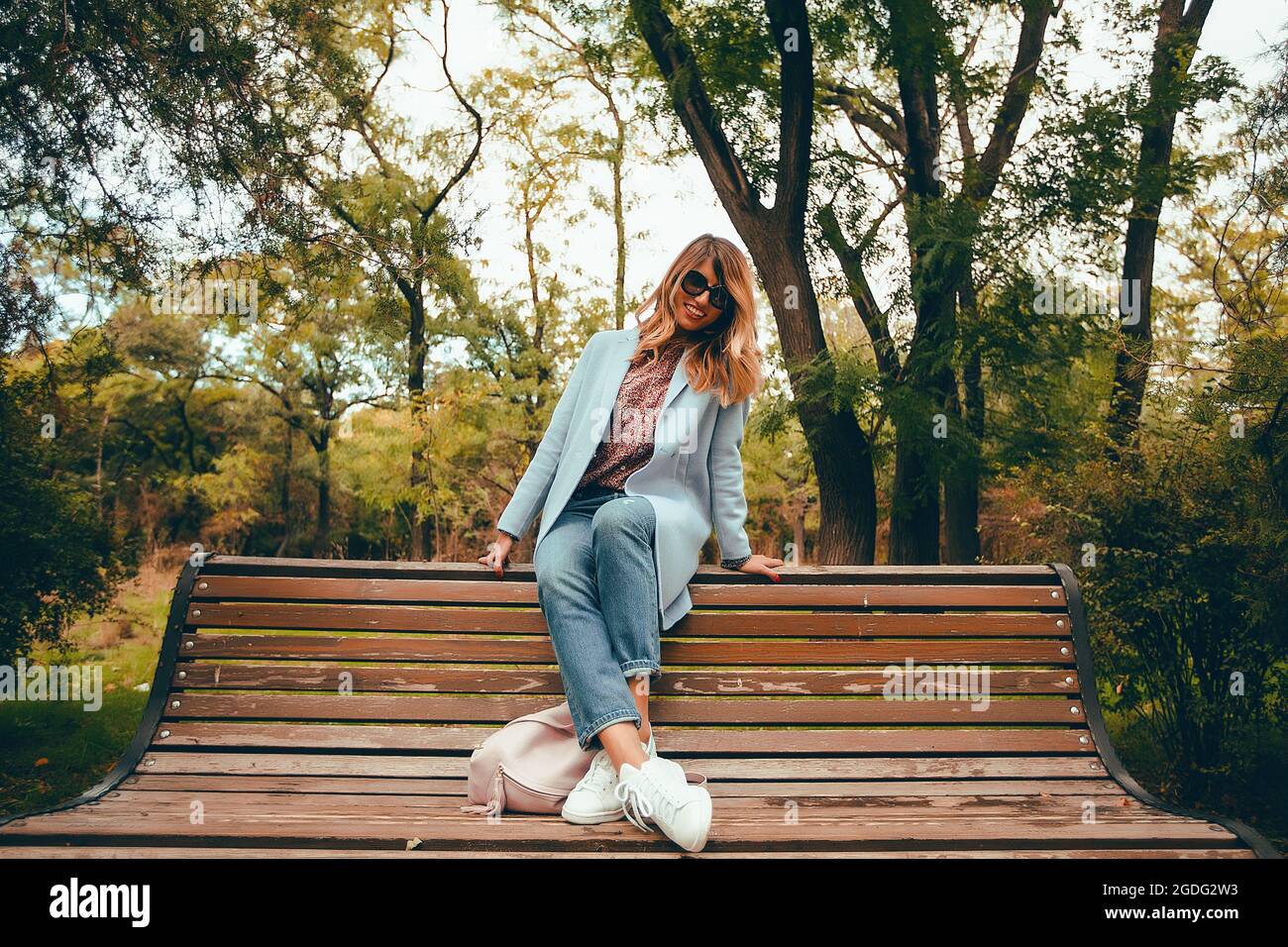 Elegante donna con capelli lunghi biondi seduta su una panchina nel parco a piena lunghezza ritratto Foto Stock