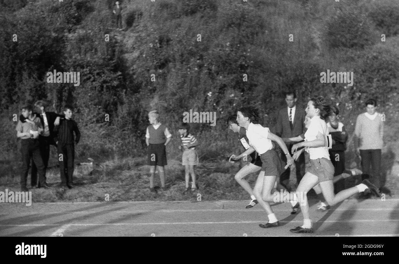 1964, storico, fuori dal comune in una giornata di sport tra le scuole della contea, le ragazze che gareggiano nella corsa di staffetta, i corridori in arrivo passano il testimone al prossimo corridore in attesa in pista, Exeter, Devon, Inghilterra, Regno Unito. Foto Stock