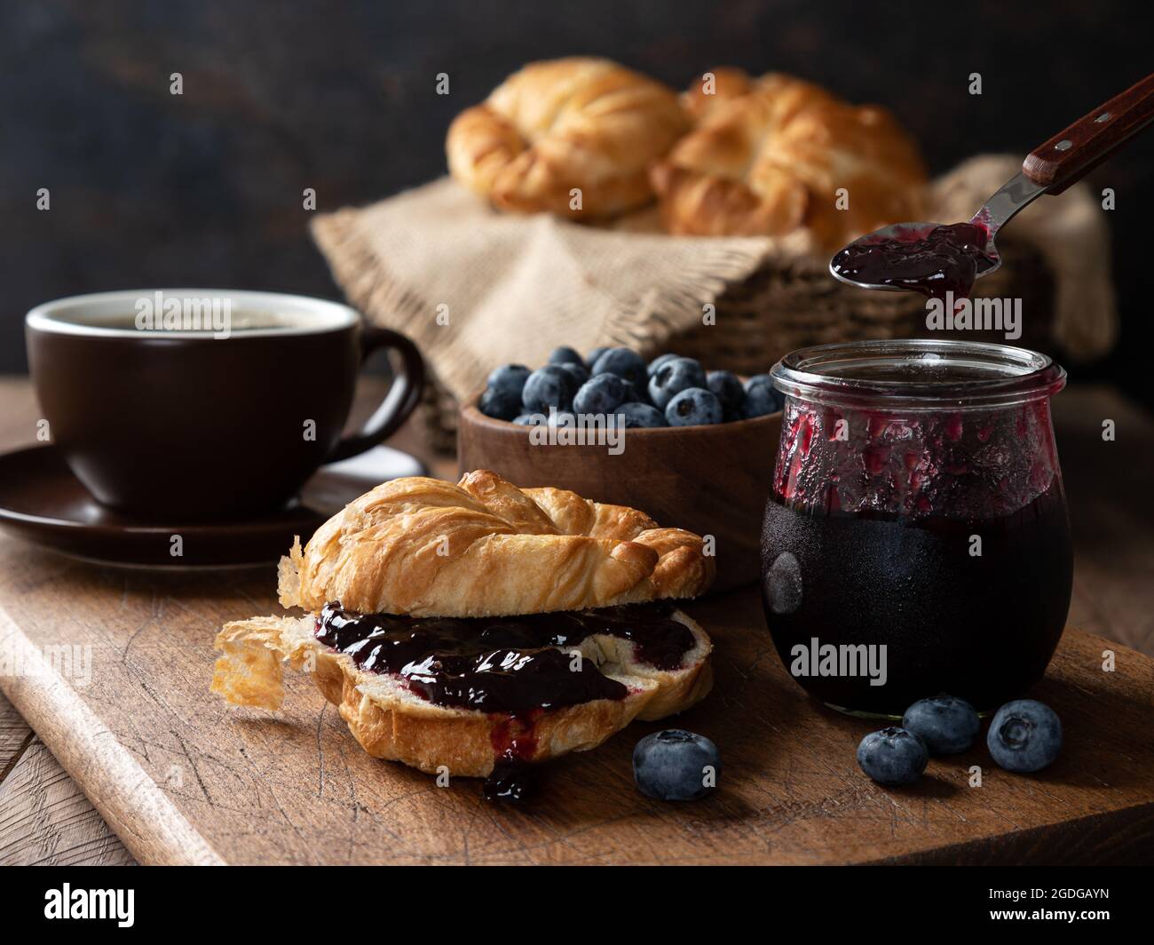 Croissant con confettura di mirtilli e mirtilli su tavola di legno con caffè, ciotola di frutti di bosco e cesto di croissant sullo sfondo Foto Stock