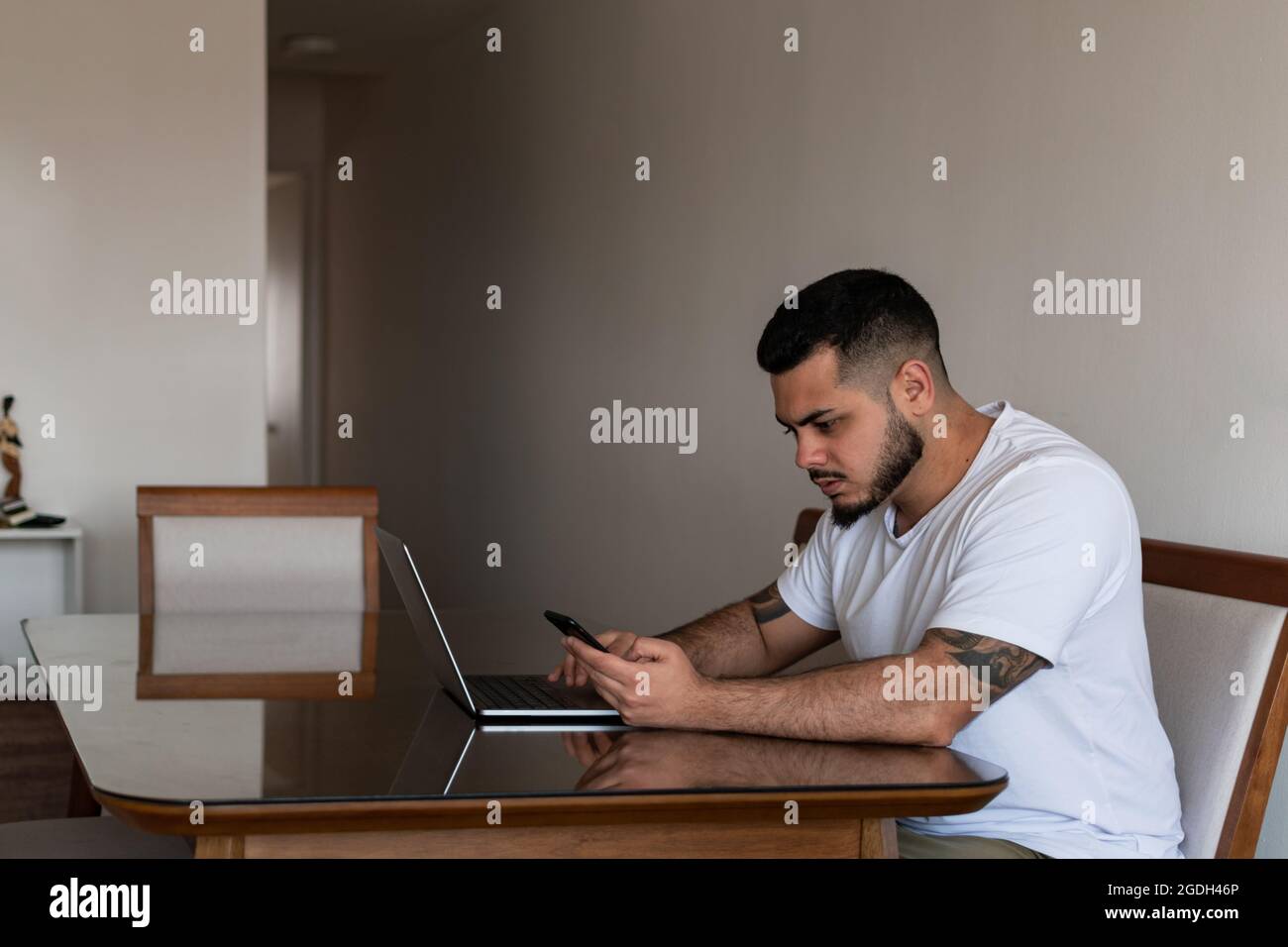 Uomo tatuato latino utilizzando cellulare e computer portatile allo stesso tempo con un volto concentrato Foto Stock