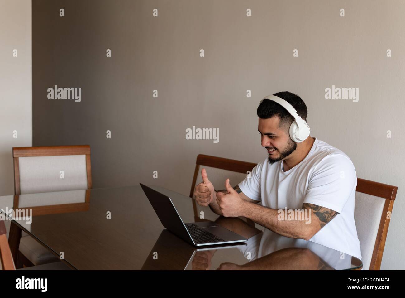 Uomo latino con tatuaggi e cuffie che mostrano i pollici fino allo schermo del computer portatile Foto Stock