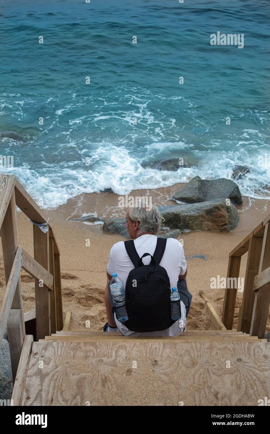 uomo maturo con capelli grigi e uno zaino seduto su una scala di legno vicino al mare Foto Stock