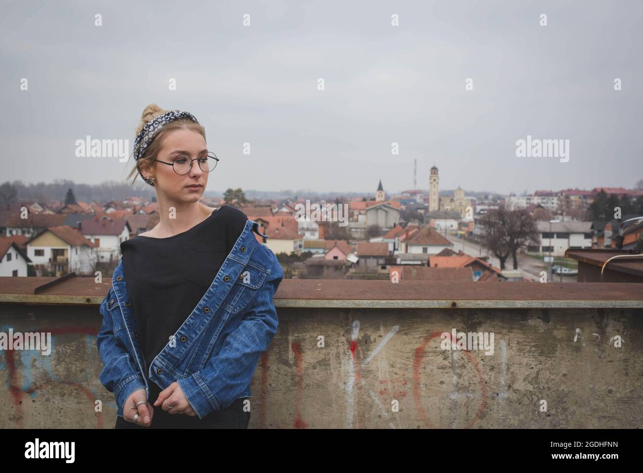 Giovane bionda multirazziale che indossa archetto, occhiali, blusa e giacca jeans in una città vecchia Foto Stock