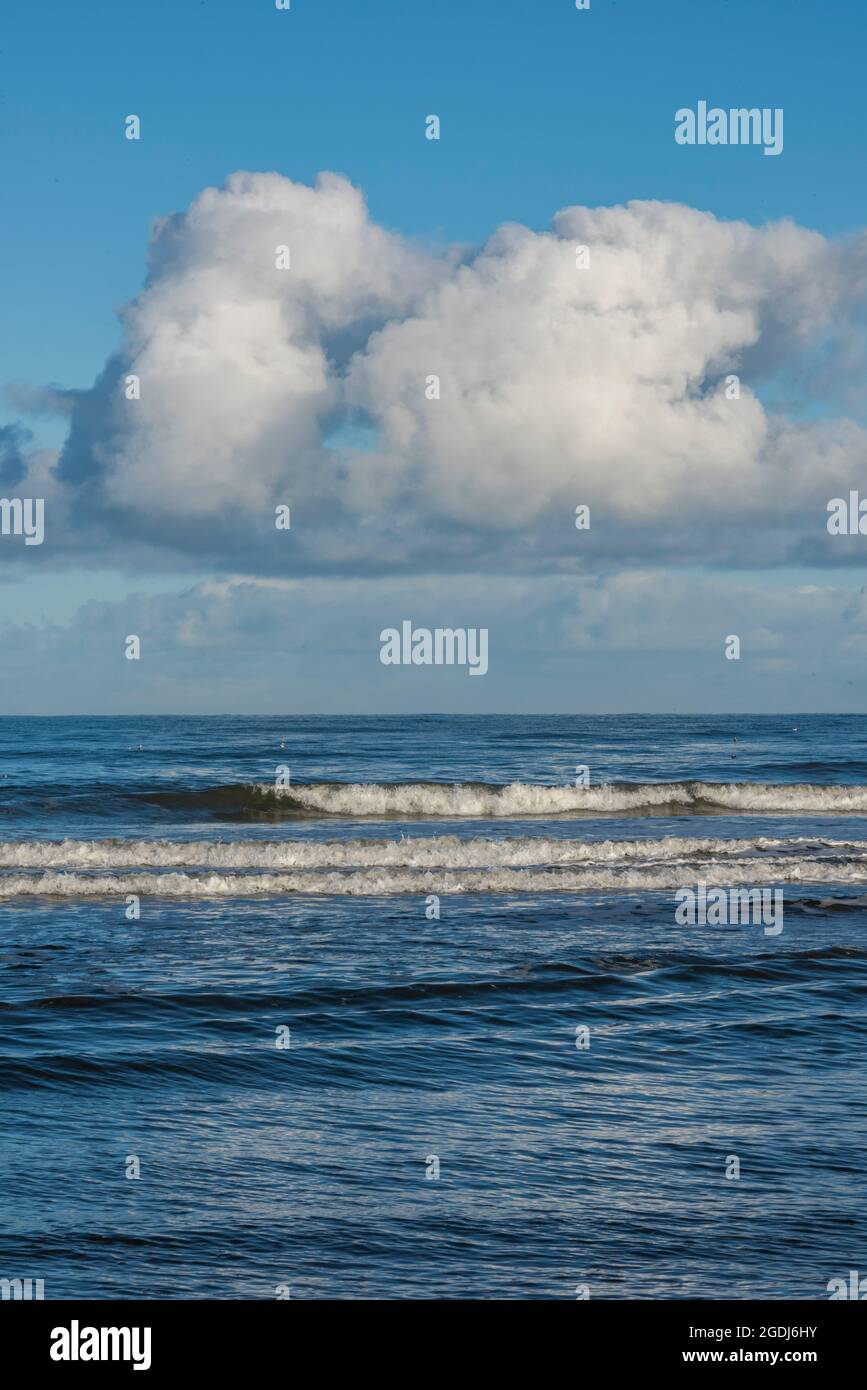onde che si infrangono su una riva remota con nuvole bianche e soffici e gli scetisti nel surf, stagni il giorno d'autunno, surf crashing sulla spiaggia in riva al mare Foto Stock