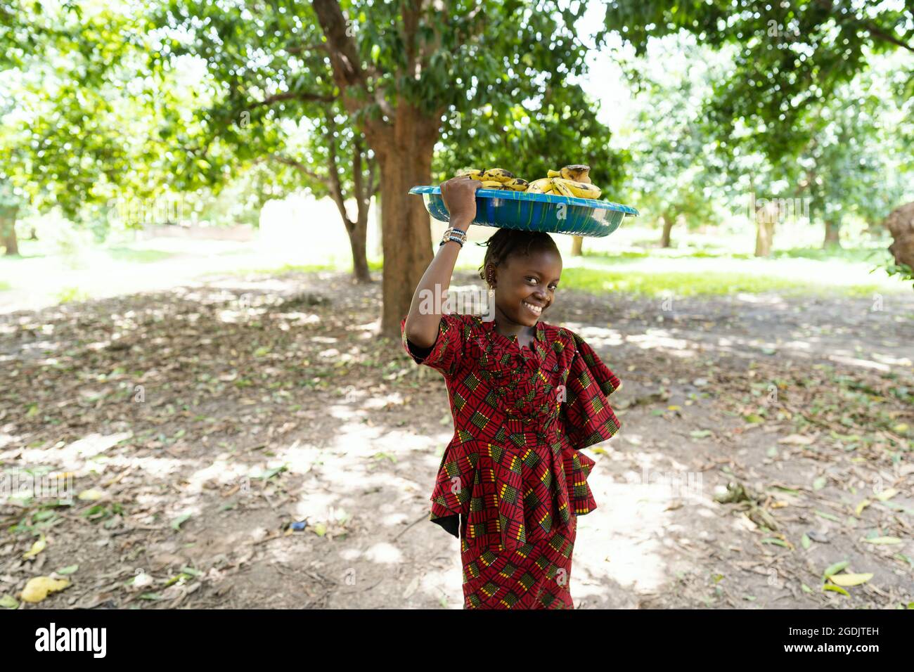 Ben vestito sorridente ragazza africana nera con un piatto di banane sulla testa, in posa per la macchina fotografica Foto Stock