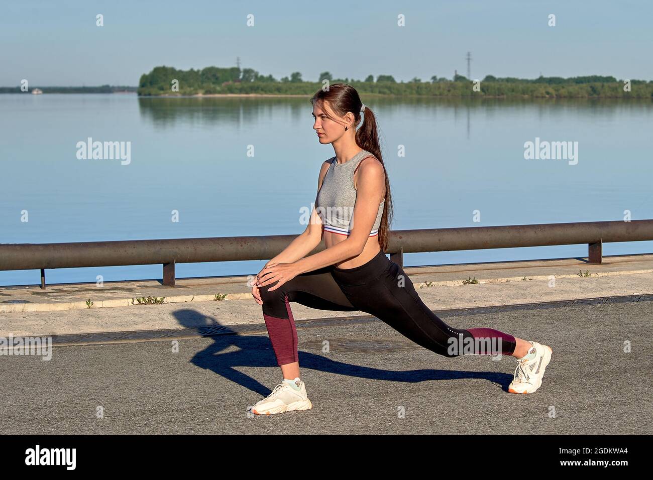 Una giovane donna fa un riscaldamento sul molo la mattina presto, sullo sfondo del fiume Foto Stock