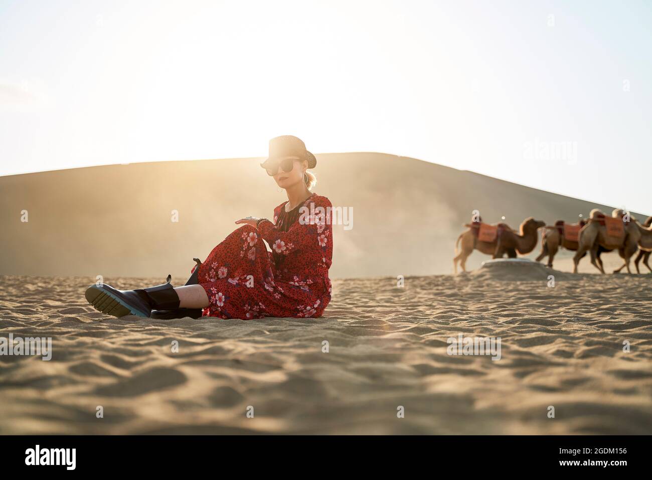 donna asiatica in abito rosso seduta nel deserto guardando la vista con caravan di cammelli e enorme duna di sabbia sullo sfondo Foto Stock