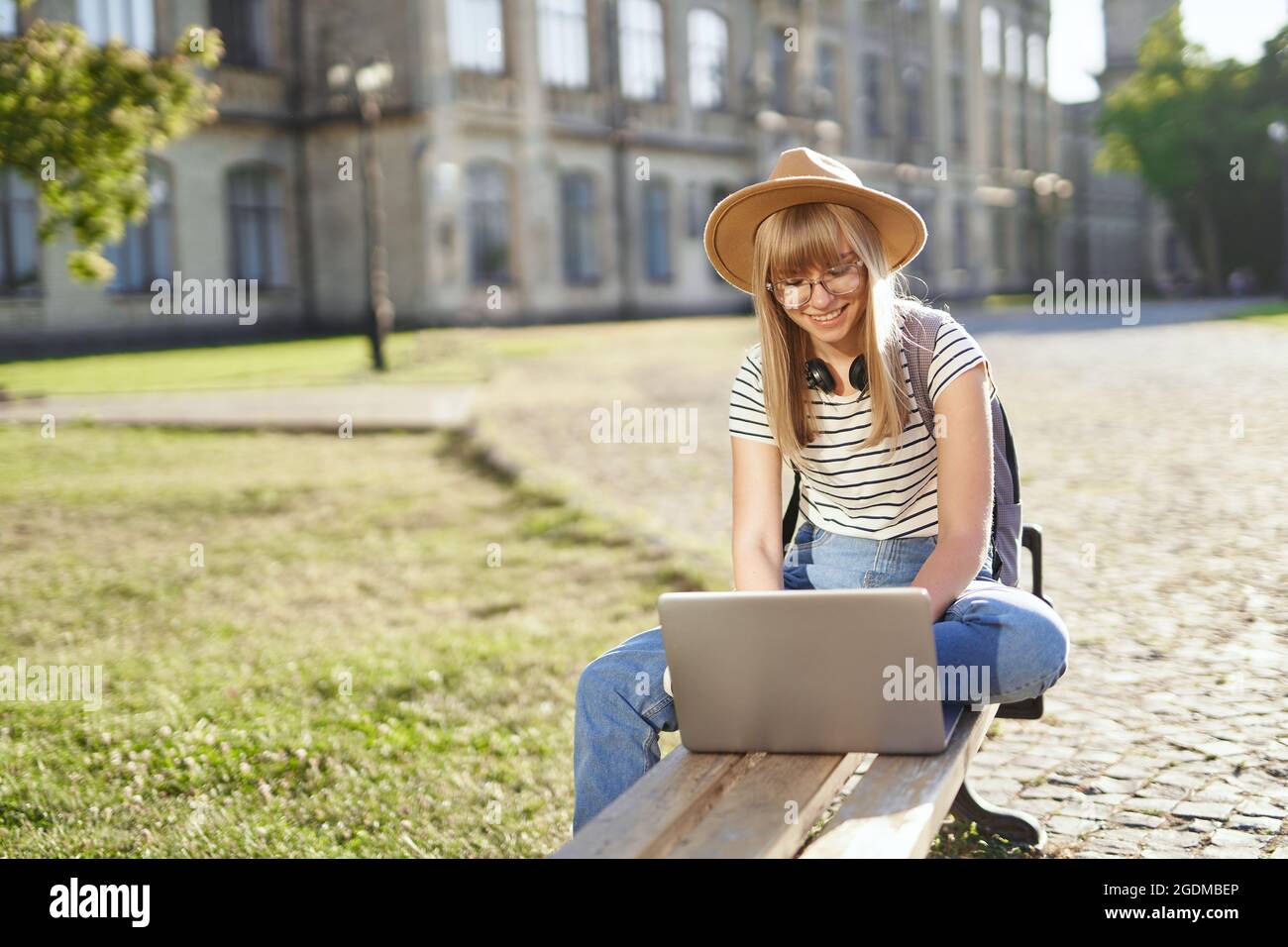 Concetto di e-learning, studio a distanza o concetto di apprendimento a distanza, giovane ragazza della scuola bionda felice, studente universitario o universitario con zaino e cappello utilizzando un computer portatile seduto sul banco del campus universitario Foto Stock