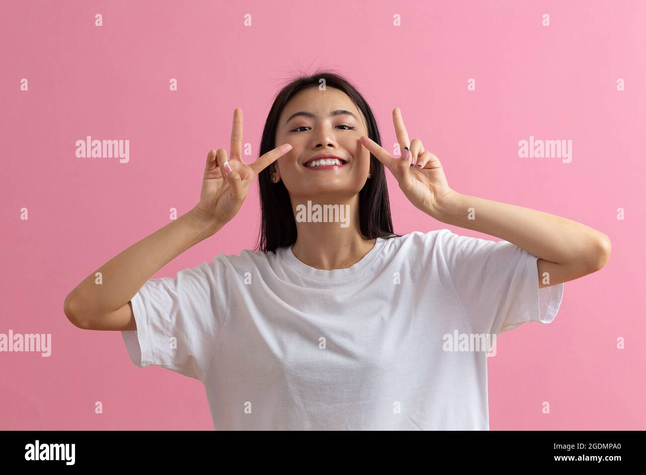 Ritratto di una donna sorridente asiatica positiva con un gesto delle dita a V su sfondo rosa. Foto Stock