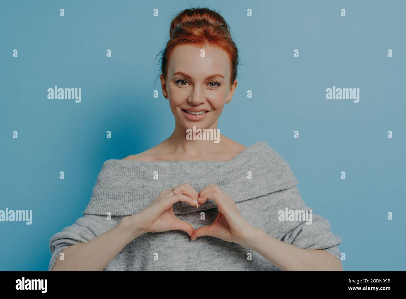 Genere donna sorridente dai capelli rossi che mostra il gesto del cuore sul petto isolato su sfondo blu della parete Foto Stock