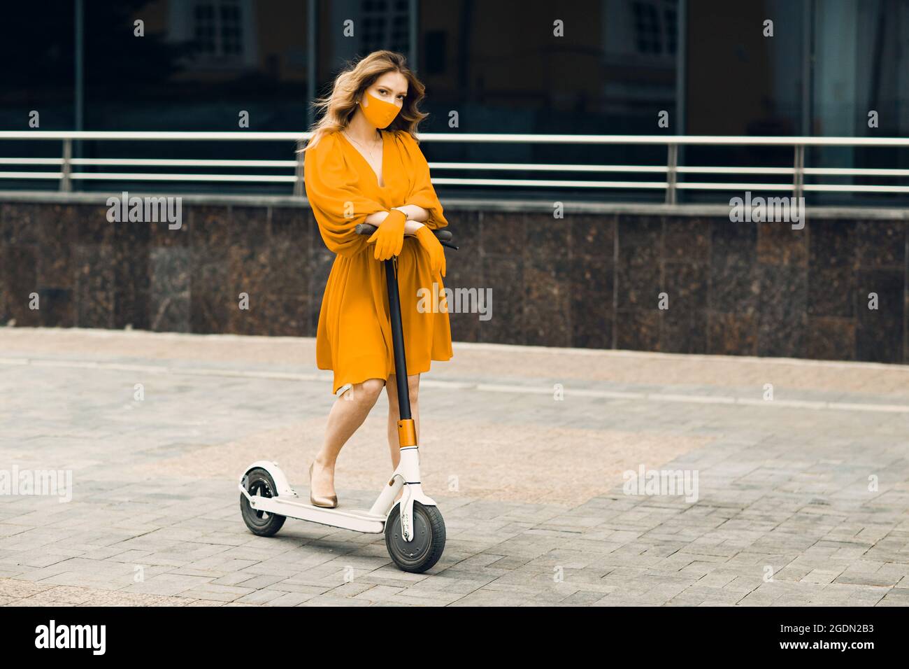 Giovane donna con scooter elettrico in abito giallo e guanti con maschera in città. Nuovo concetto di protezione COVID-19 di modo normale e coronavirus Foto Stock