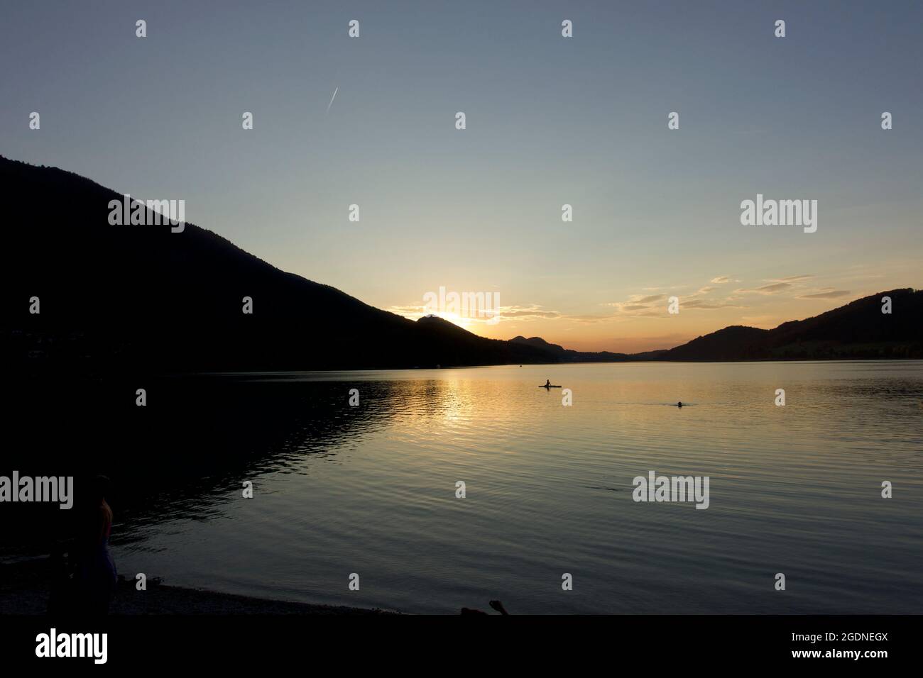 Lago tranquillo con due nuotatori solitari a fine estate. Cielo blu e sole tramontato che scende dietro le montagne, in silhouette, lago Fuschl, Austria Foto Stock