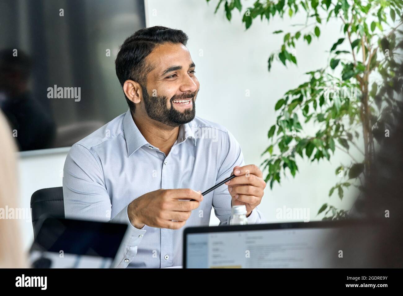 Sorridente CEO indiano di sesso maschile che ascolta collega tenendo la penna in mani al tavolo. Foto Stock