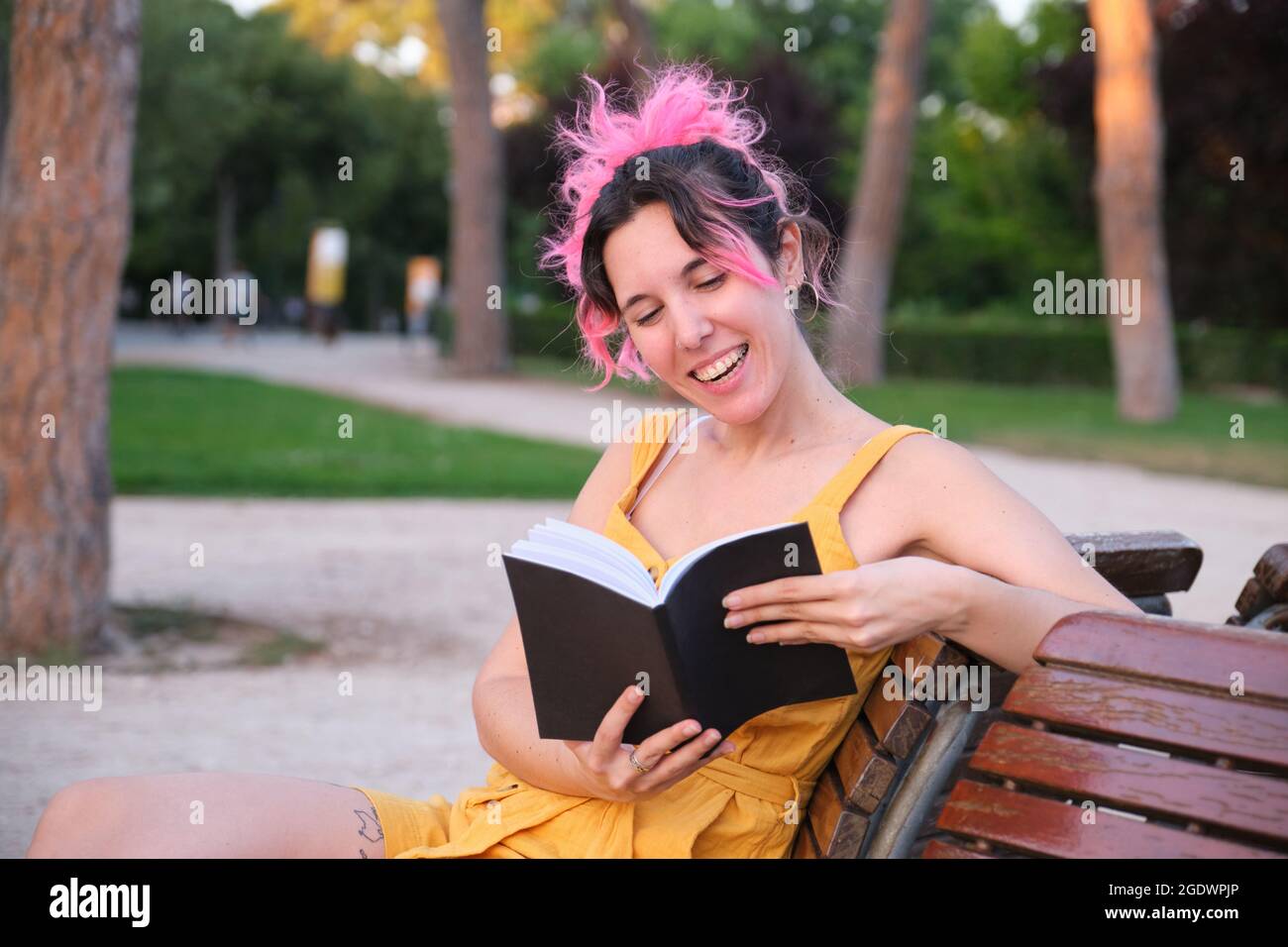 Giovane donna caucasica con capelli rosa seduta in panca e lettura di un libro. Foto Stock