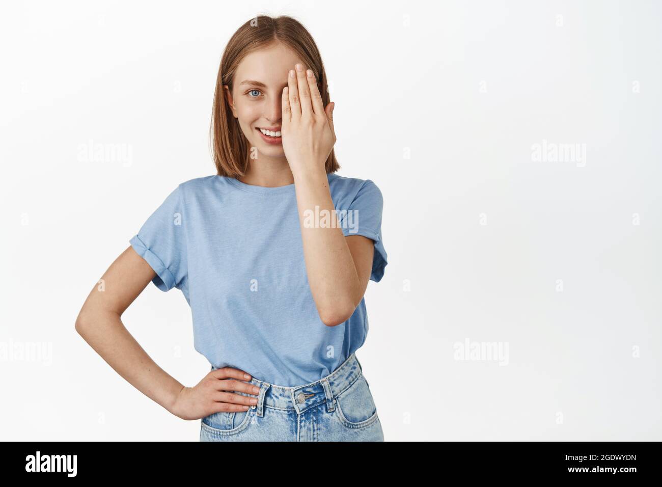 Bella donna bionda coprire metà del viso, guardando con un occhio e sorridente sicuro, prima e dopo l'effetto, in piedi in t-shirt blu contro bianco Foto Stock