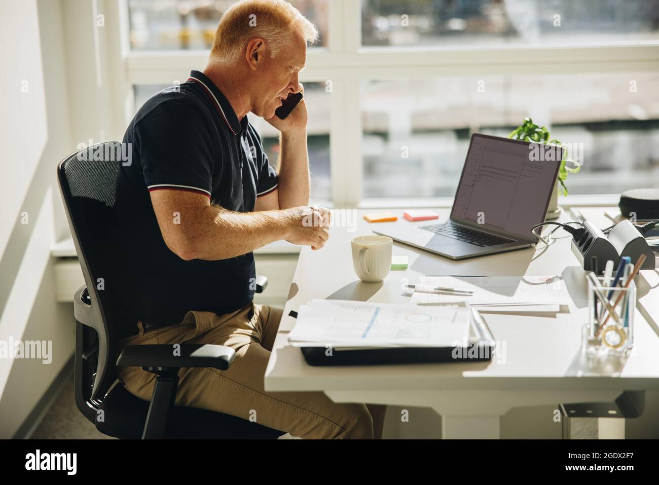 uomo d'affari che parla al telefono cellulare mentre è seduto di fronte al laptop. Uomo anziano seduto alla sua scrivania a fare una telefonata. Foto Stock