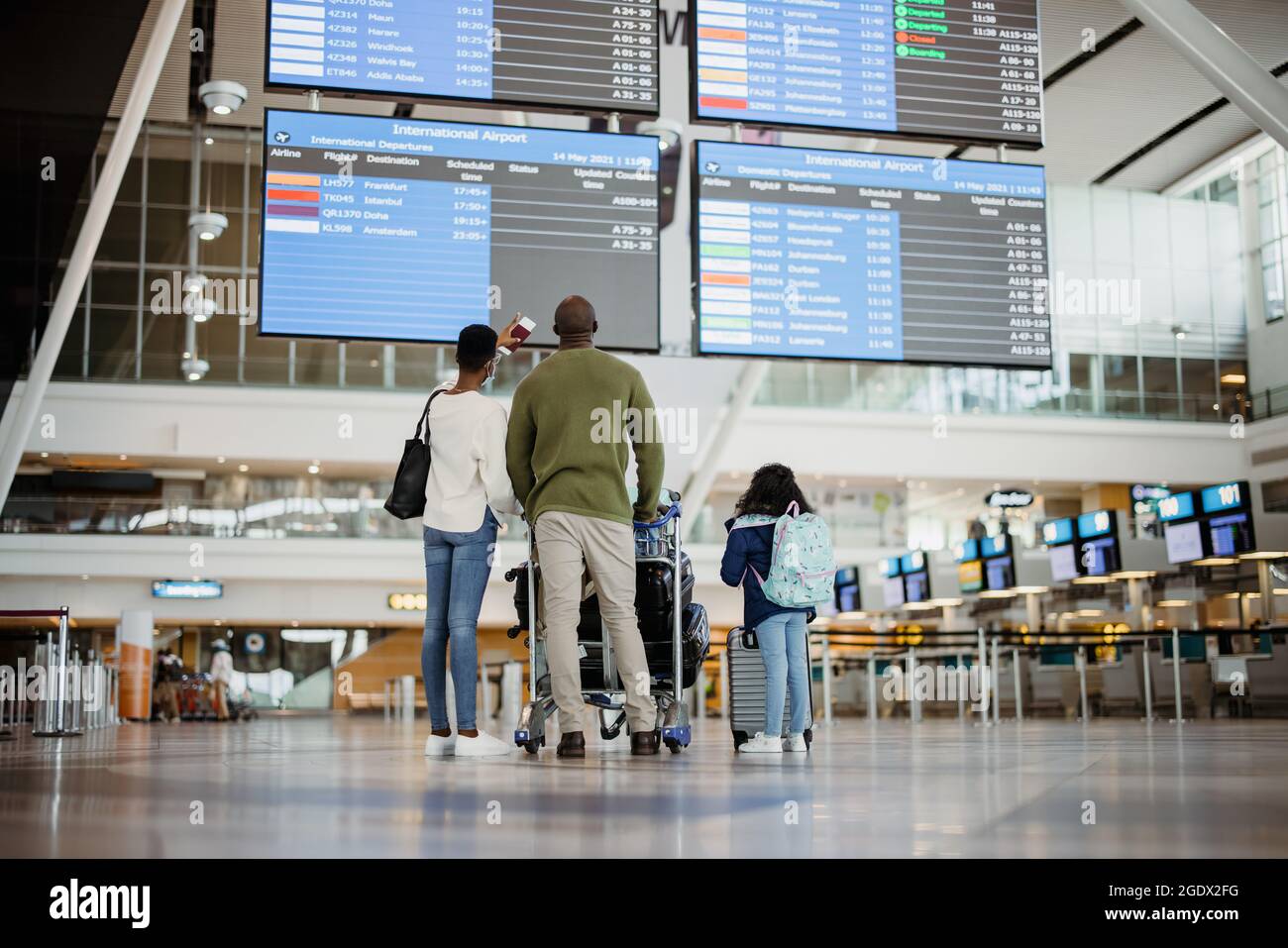 Vista posteriore della famiglia africana con bagagli che guardano al tavolo orario dei voli di linea al terminal dell'aeroporto. Genitori con figlia in attesa all'aeroporto e che Foto Stock