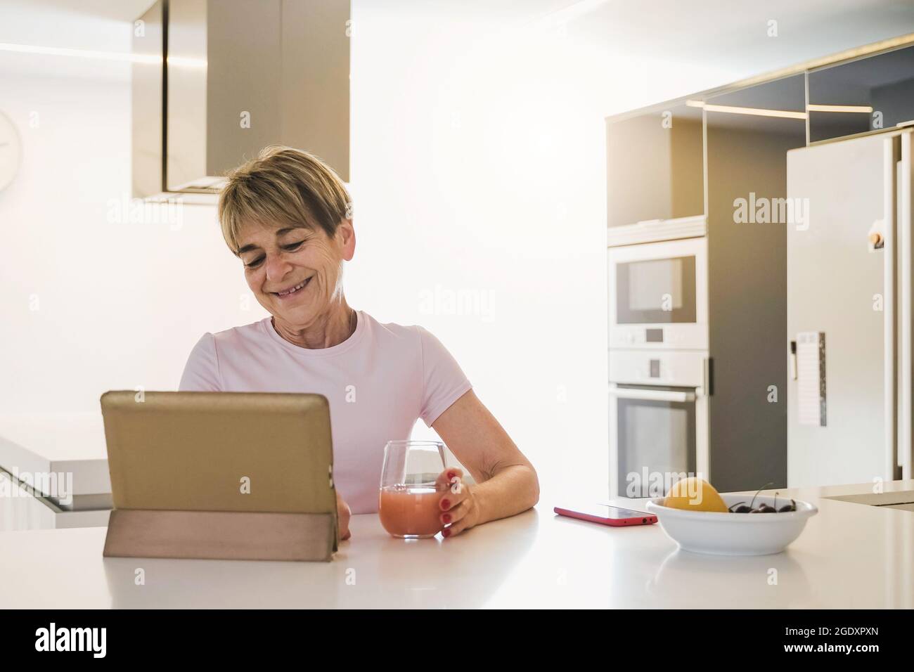 Donna anziana felice che usa il tablet digitale mentre si mangia la colazione in cucina - Focus on Face Foto Stock