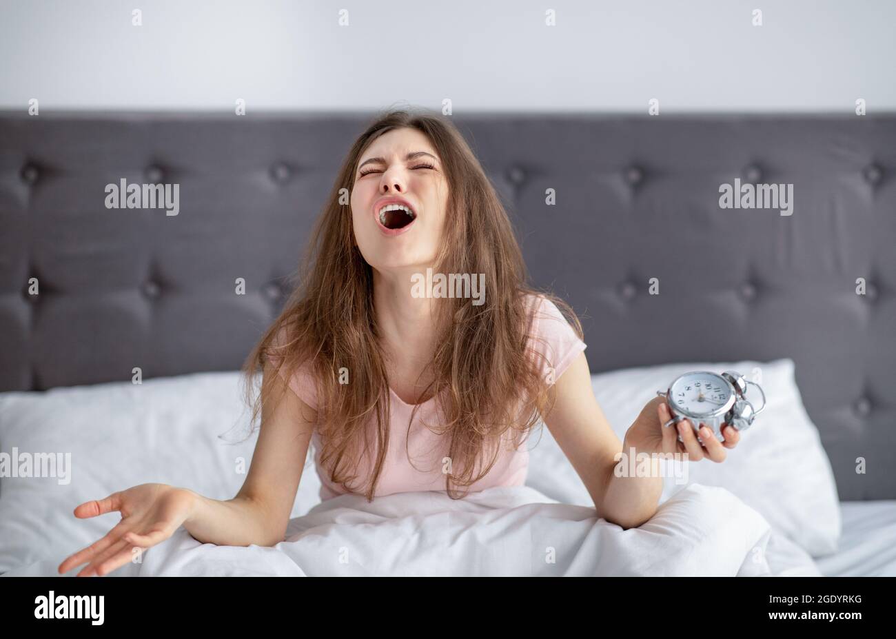 Donna giovane disperata con sveglia seduta a letto, sentendosi stressata al mattino, non disposta a mettersi al lavoro a casa Foto Stock