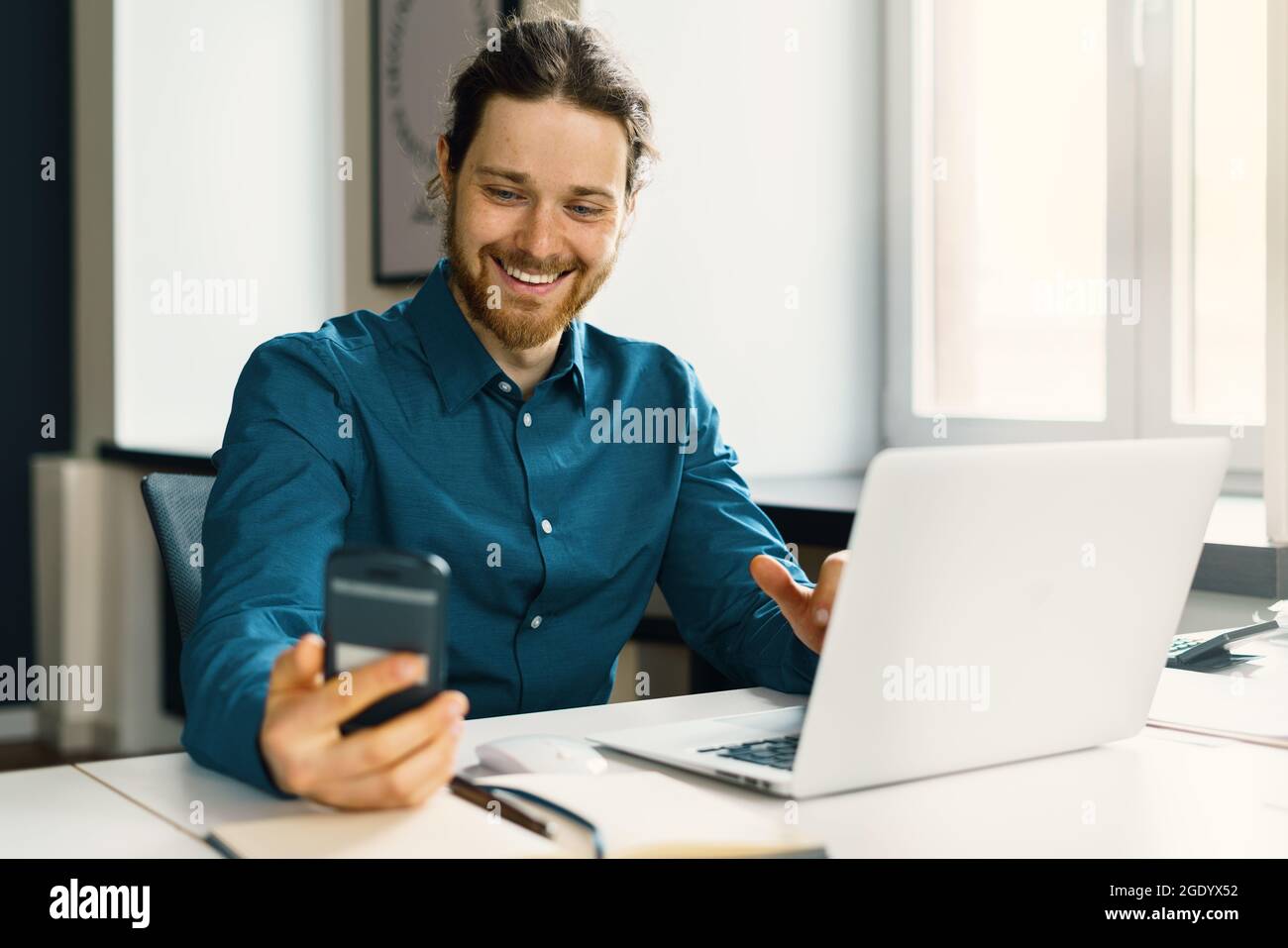 Un giovane sorridente in un abbigliamento casual che guarda lo schermo dello smartphone sul posto di lavoro Foto Stock