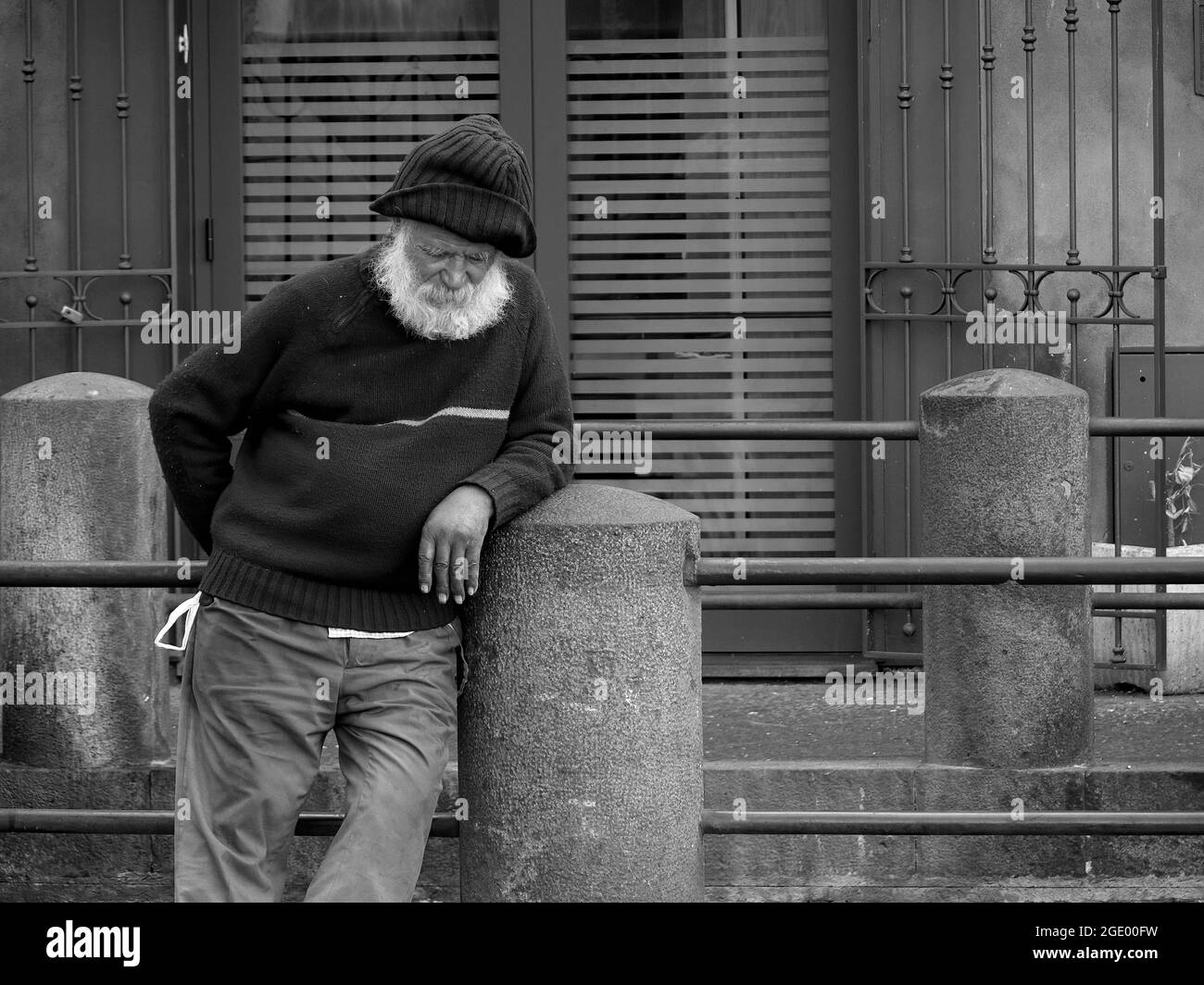 Un vecchio trampino con barba bianca è appoggiato alla colonna di pietra di un ringhiera che snooze nel centro storico di Catania, in Sicilia Foto Stock