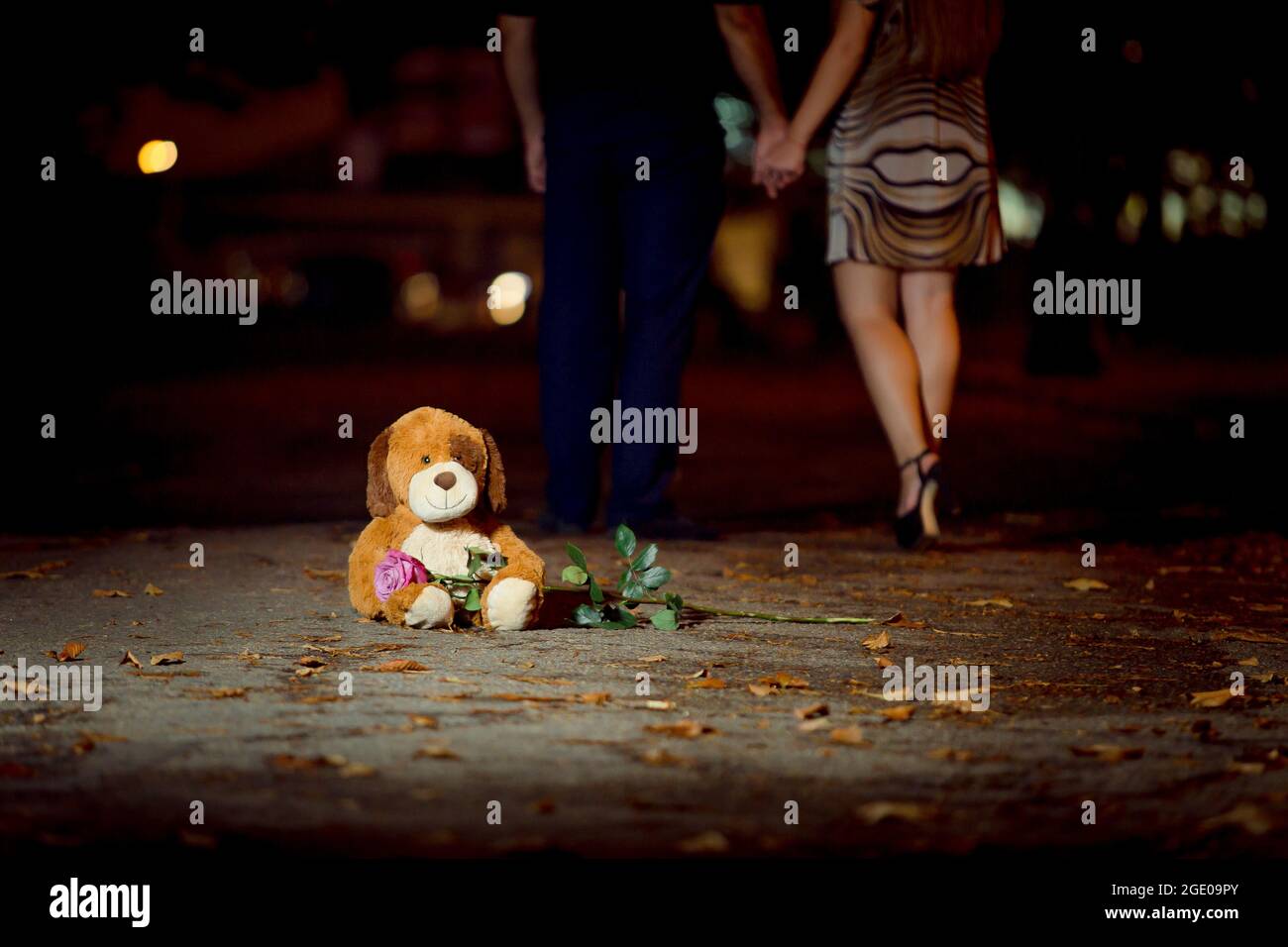 Un orsacchiotto si siede sull'asfalto e tiene una rosa fresca. Sullo sfondo delle gambe di una coppia amorevole di uomo e donna. Foto Stock