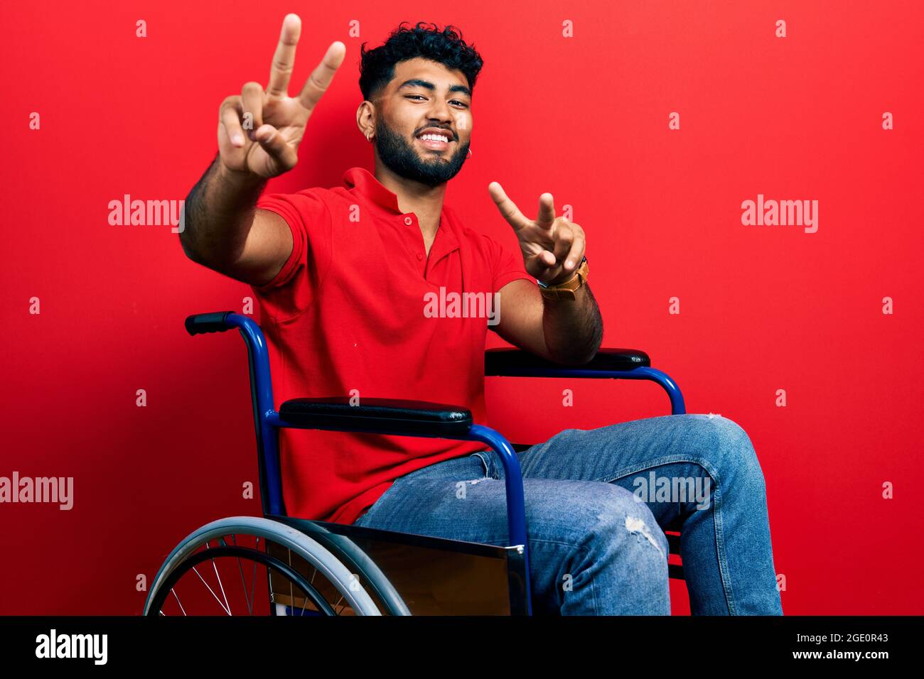 Uomo arabo con barba seduta su sedia a rotelle che sorride con la lingua che mostra le dita di entrambe le mani che fanno il segno della vittoria. Numero due. Foto Stock
