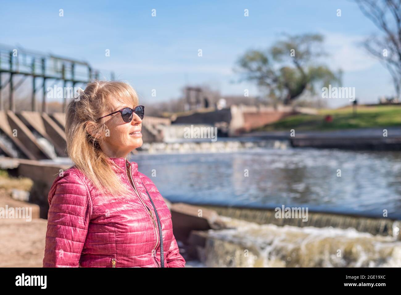 Vista di un'allegra donna adulta che guarda lontano con occhiali da sole in piedi nel parco con un laghetto dietro in una giornata di sole. Foto Stock