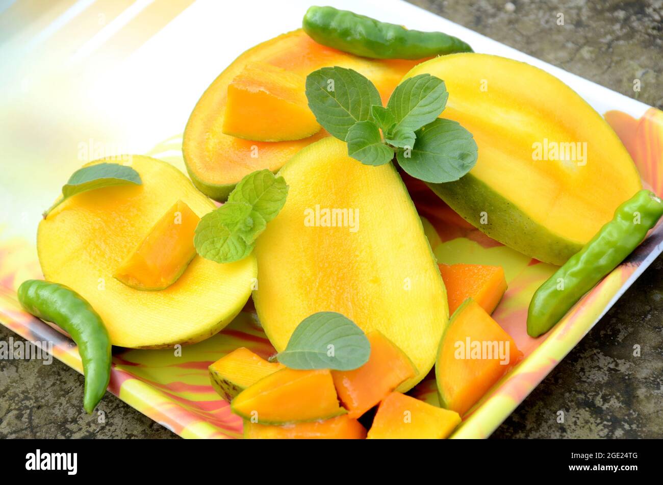 chiuda la frutta di mango giallo verde matura tagliata a fette con le foglie di menta verde e fredda nel vassoio sopra fuori di fuoco sfondo marrone grigio. Foto Stock