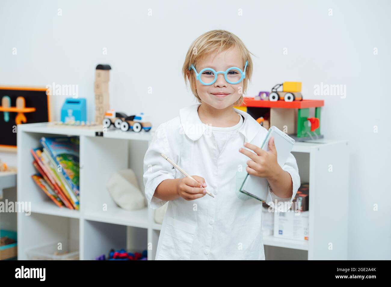 Ritratto di un ragazzino con un sorriso gioioso che suona un medico. Indossa occhiali giocattolo e un abito bianco, tenendo un taccuino e una matita. A un bambino l Foto Stock